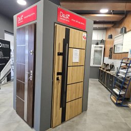 TECH-DOM S.C. JAROSŁAW BUSZKA DARIUSZ DARGACZ - Ekspozycja dwóch modeli drzwi zewnętrznych Delta na szarym postumencie w salonie, z widocznymi detalami jak klamki, zamki i oznaczenia producenta, na tle elementów wyposażenia wnętrza.