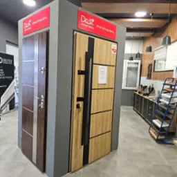 Ekspozycja dwóch modeli drzwi zewnętrznych Delta na szarym postumencie w salonie, z widocznymi detalami jak klamki, zamki i oznaczenia producenta, na tle elementów wyposażenia wnętrza.