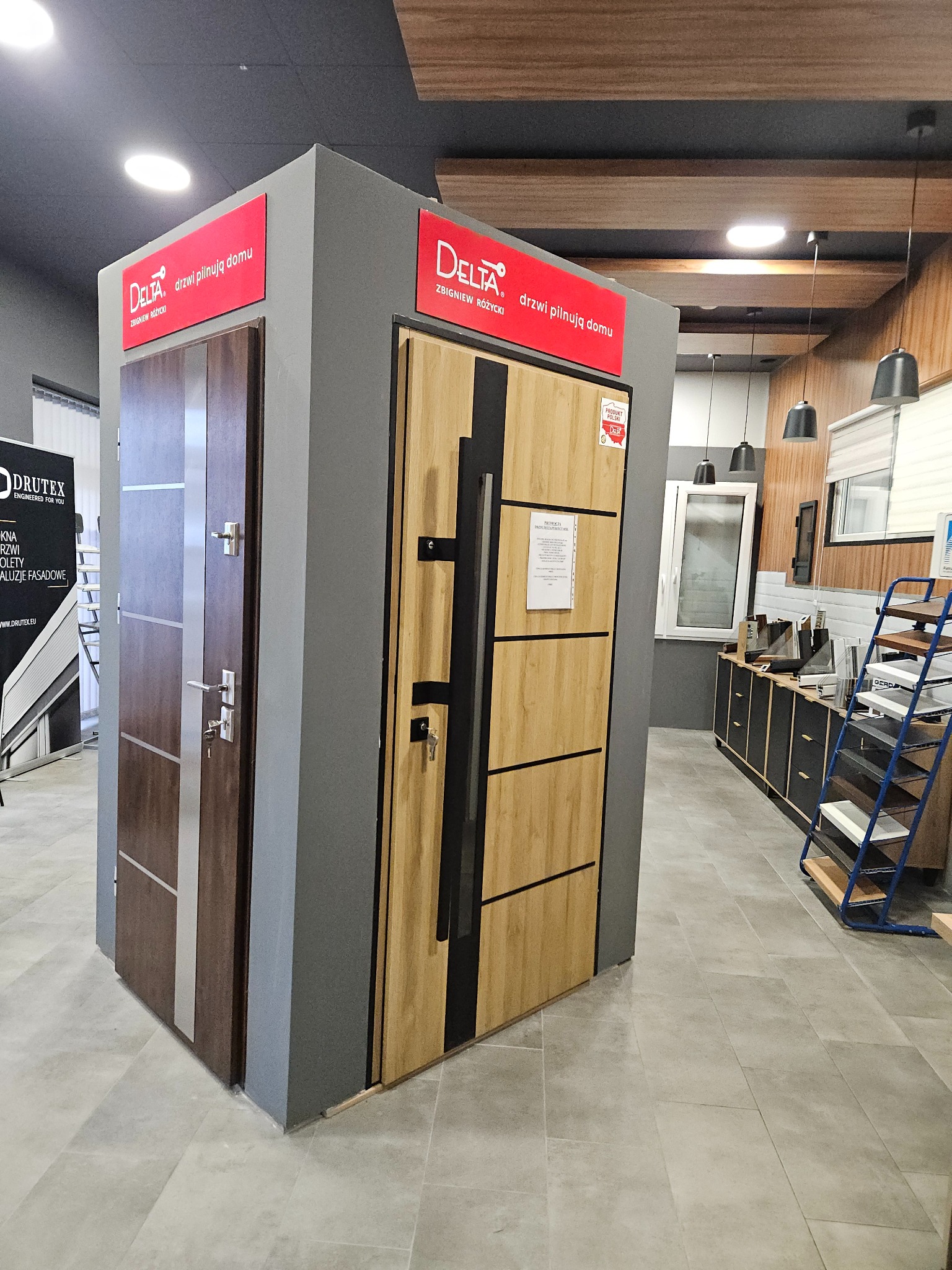 Ekspozycja dwóch modeli drzwi zewnętrznych Delta na szarym postumencie w salonie, z widocznymi detalami jak klamki, zamki i oznaczenia producenta, na tle elementów wyposażenia wnętrza.