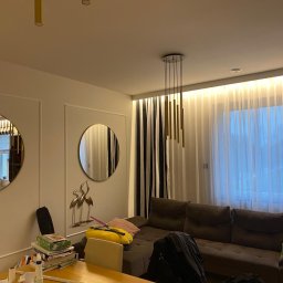 Adrian Rak - Jasne wnętrze salonu z okrągłymi lustrami w złotych ramach, brązową sofą i lampą wiszącą z złotych tub, oświetlone listwą LED pod sufitem.