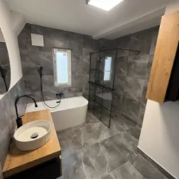 Nowoczesna aranżacja łazienki w Legnicy z szarymi płytkami imitującymi marmur, kabiną prysznicową typu walk-in z czarnymi profilami, wolnostojącą białą wanną i umywalką nablatową na drewnianym blacie.