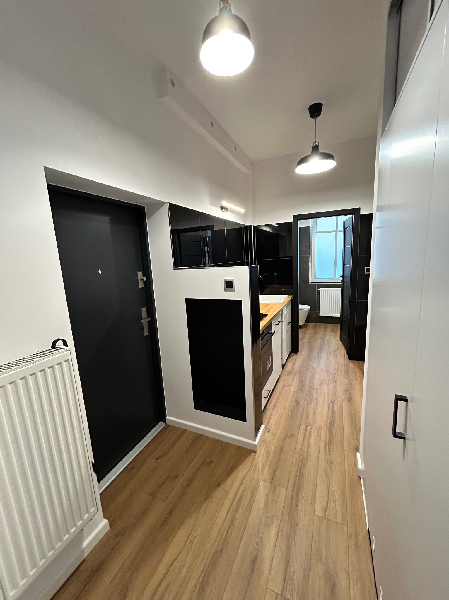 Wnętrze mieszkania po remoncie: przedpokój z zabudową kuchenną, widok na łazienkę, drewnopodobne panele na podłodze, białe ściany, czarne akcenty meblowe i drzwi.