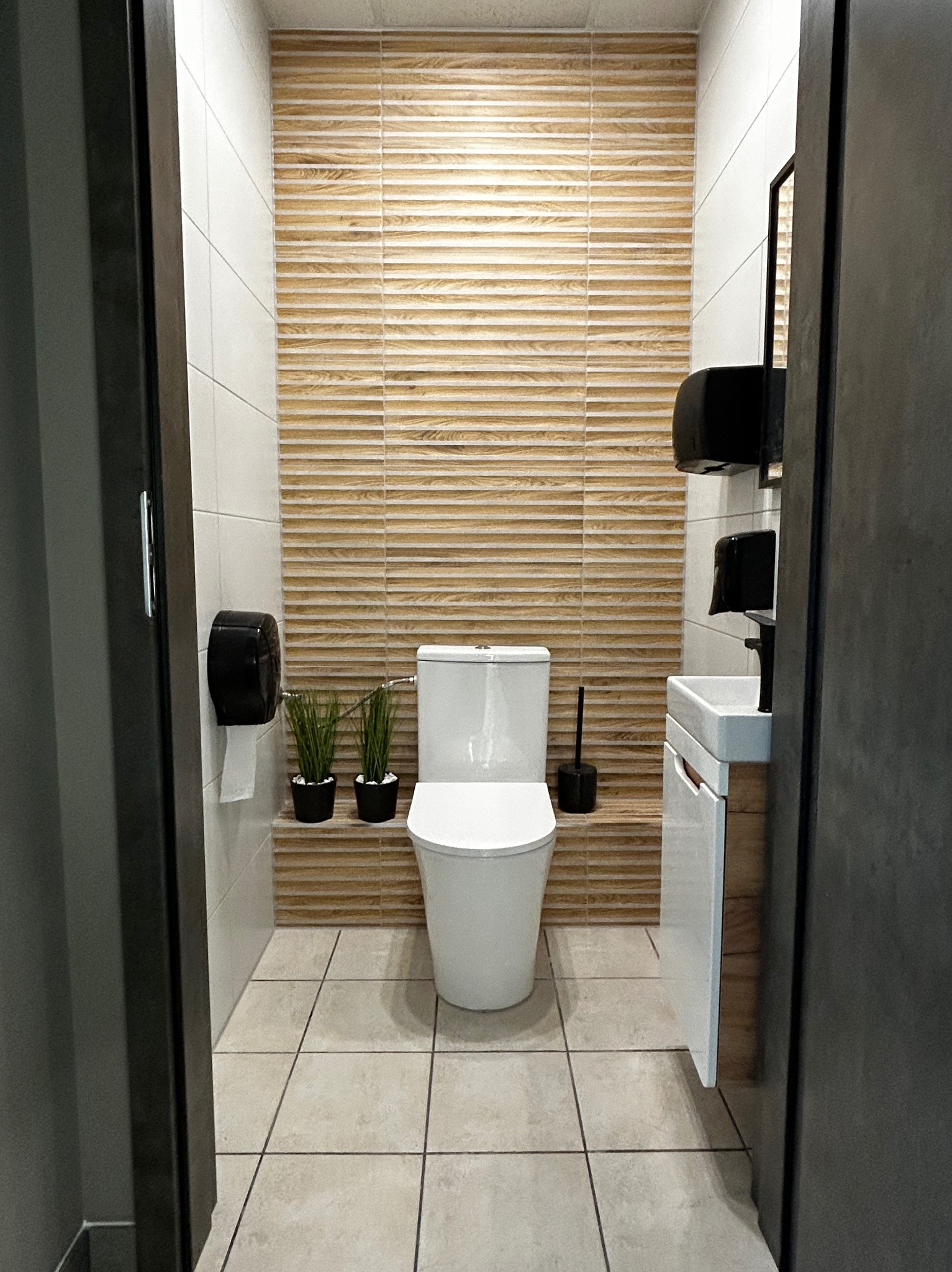 Nowoczesna łazienka z beżowymi płytkami imitującymi drewno na ścianie, białą ceramiką i minimalistyczną umywalką z szafką, widziana zza uchylonych, ciemnych drzwi.