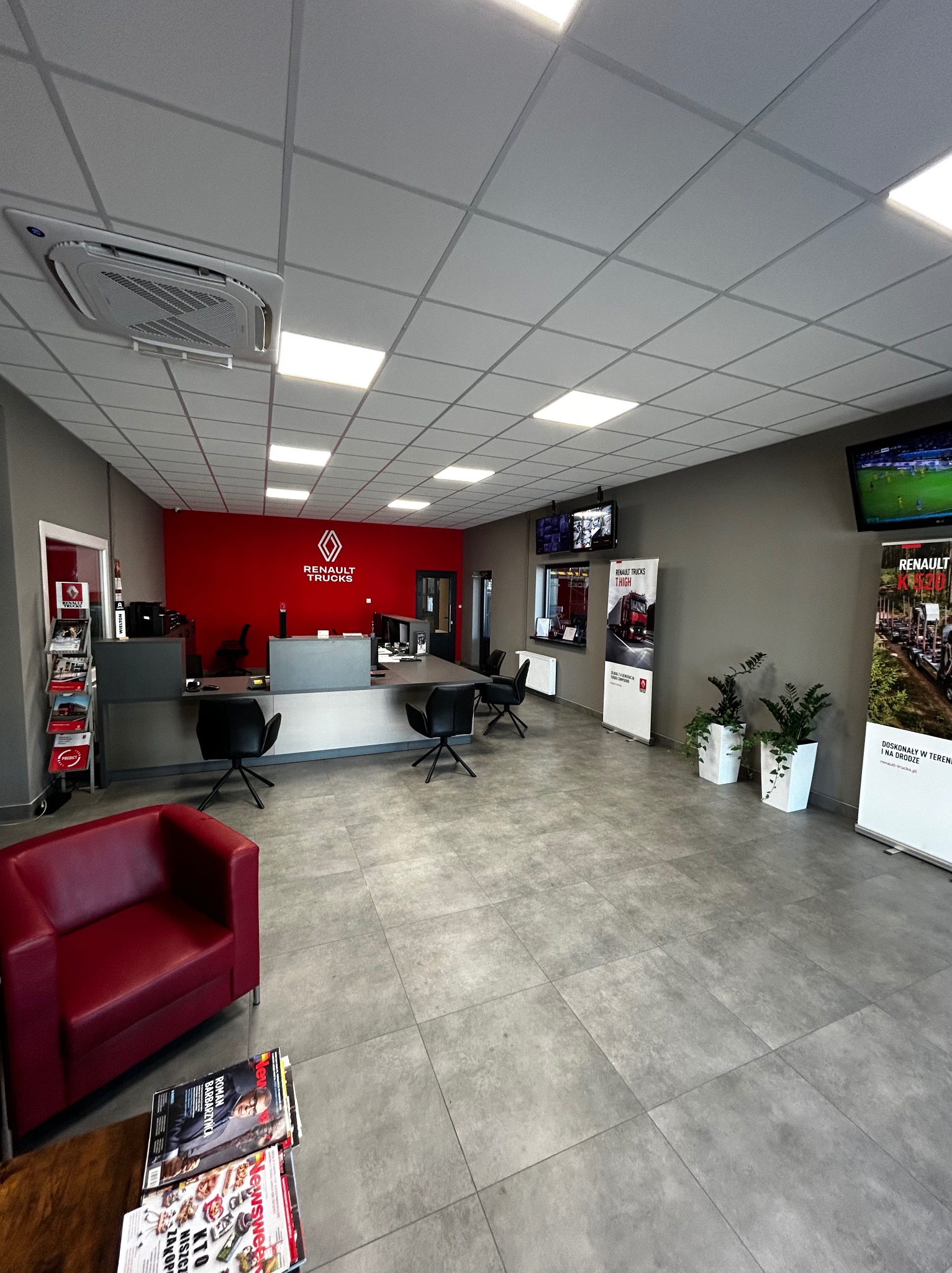 Nowoczesne wnętrze salonu samochodowego Renault Trucks z szarą podłogą z płytek, czerwonym akcentem na ścianie z logo, recepcją, czarnymi krzesłami, roślinami w białych donicach i telewizorami...