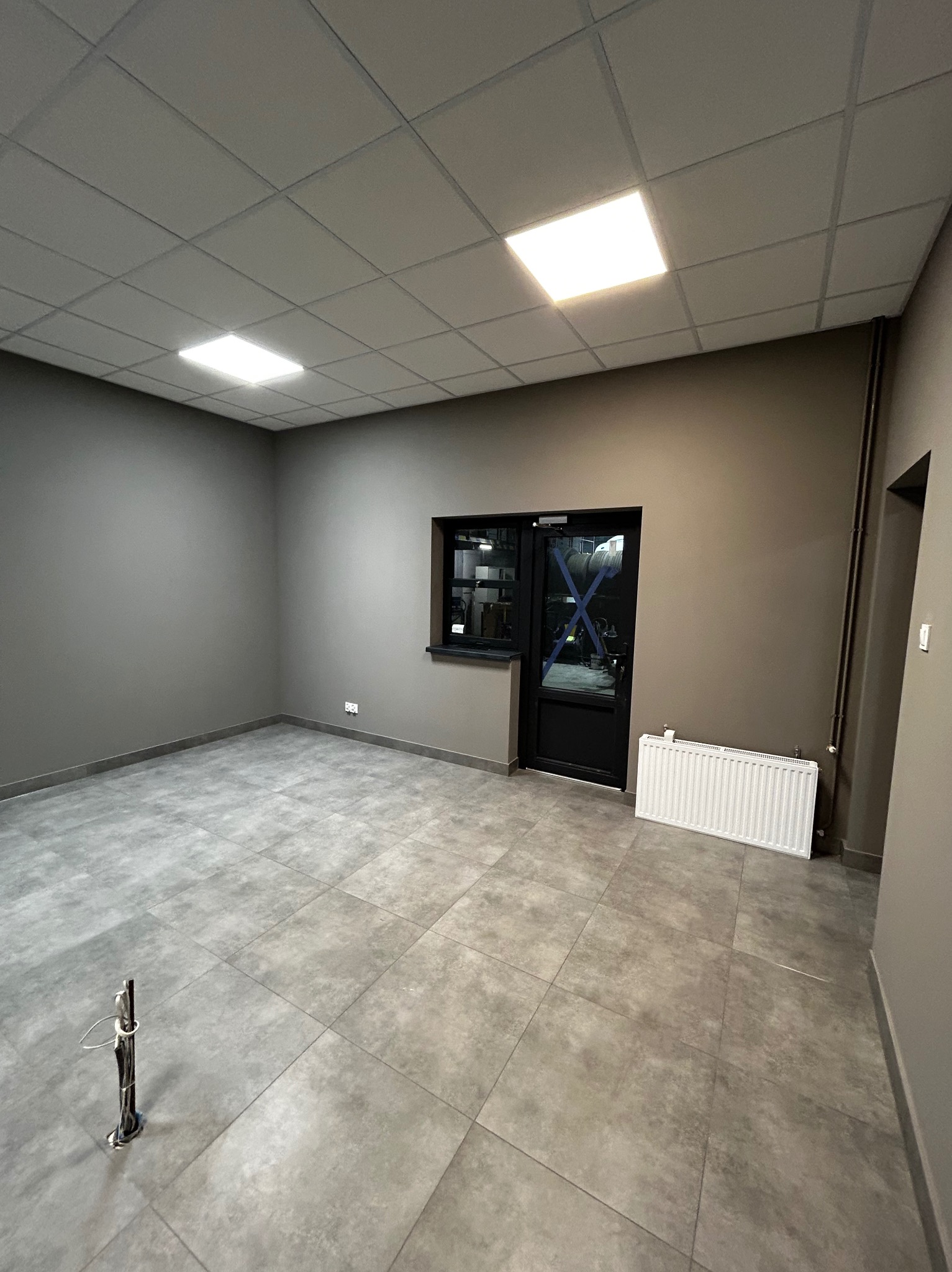 Pusty pokój z szarą podłogą z płytek i szarymi ścianami, widoczny grzejnik pod oknem i zabezpieczone taśmą okno/drzwi.