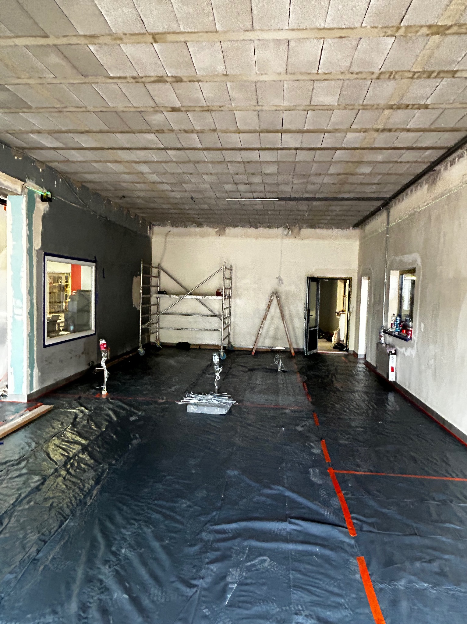 Wnętrze w trakcie prac przygotowawczych: podłoga zabezpieczona czarną folią z wyznaczonymi czerwonymi liniami, ściany częściowo wykończone, widoczne rusztowania i drabina.