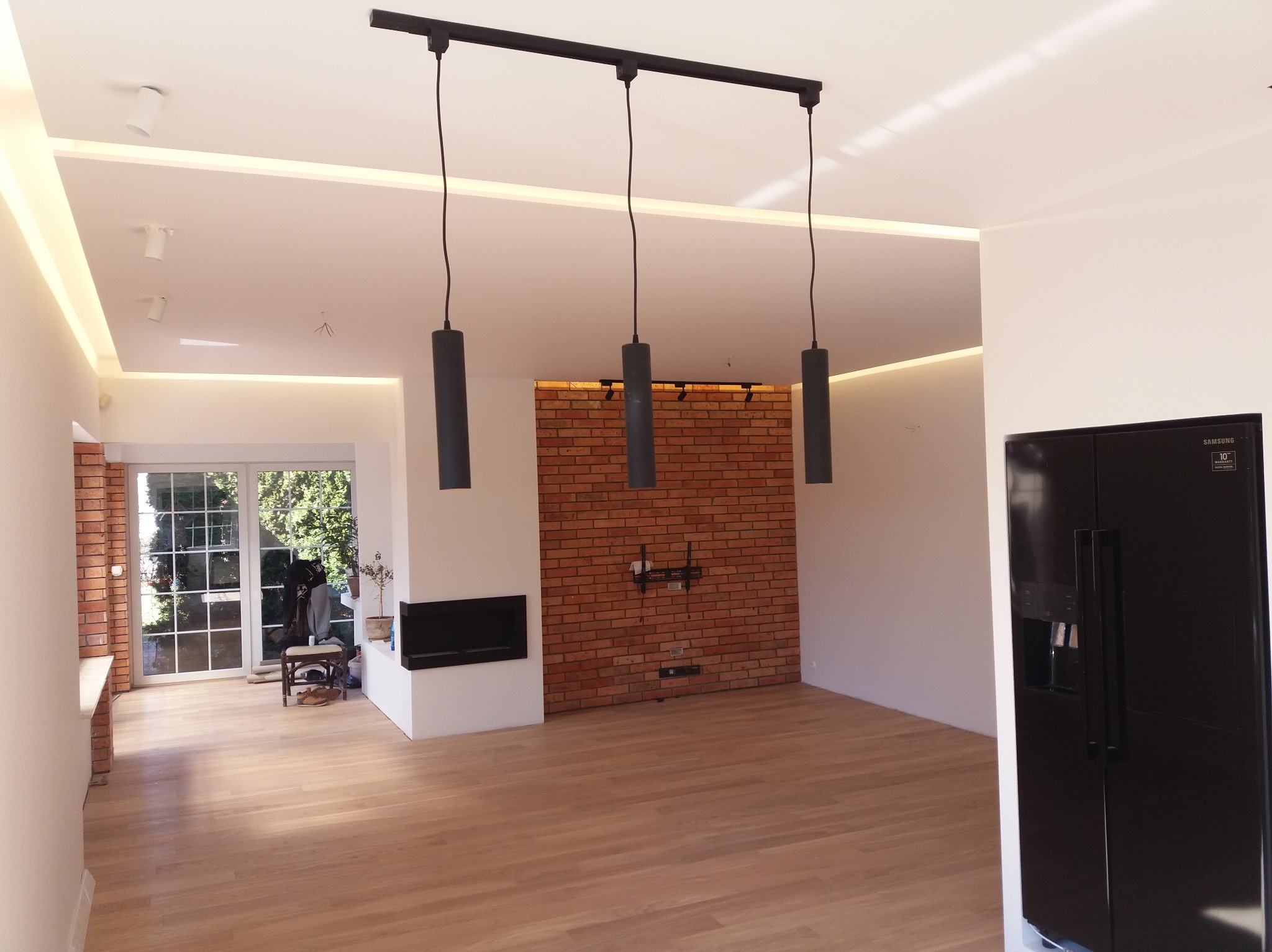 Jasne, przestronne wnętrze po remoncie z parkietem, ceglaną ścianą z instalacją pod telewizor, nowoczesnym oświetleniem sufitowym i czarną lodówką Samsung.