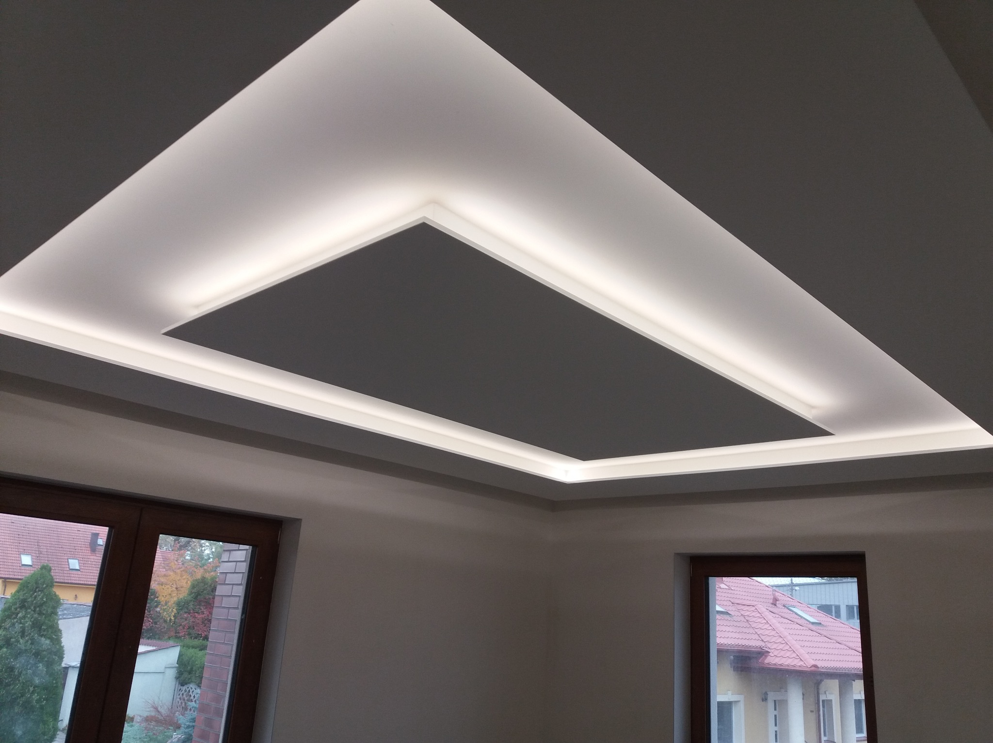 Sufit podwieszany z oświetleniem LED w kształcie prostokąta, widok na dwa okna z widokiem na jesienny ogród i sąsiednie budynki.