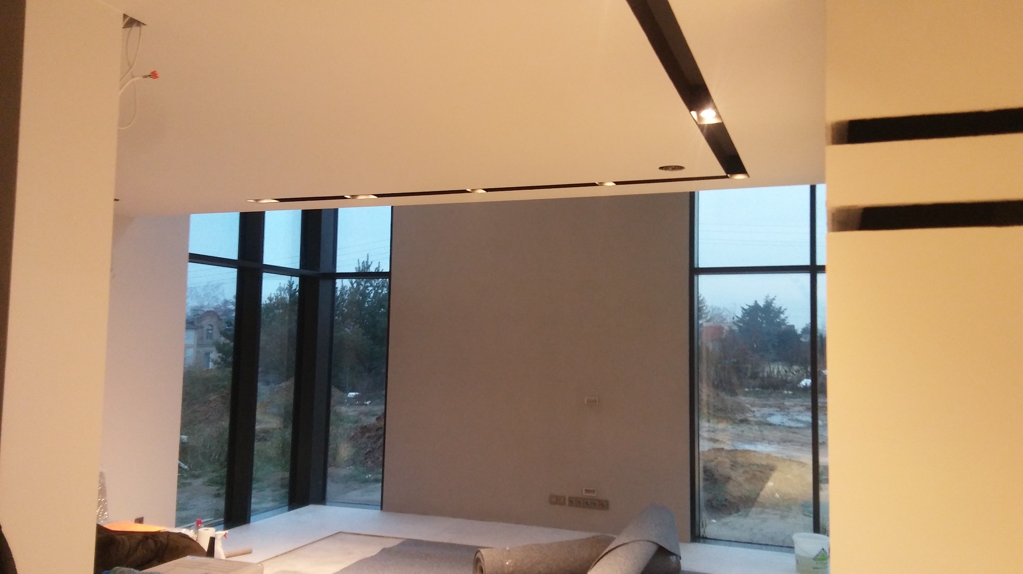 Wnętrze w trakcie wykończenia: szara ściana, duże okna z czarnymi ramami, podwieszany sufit z oświetleniem punktowym, zwinięty dywan na podłodze, widoczne przewody elektryczne.