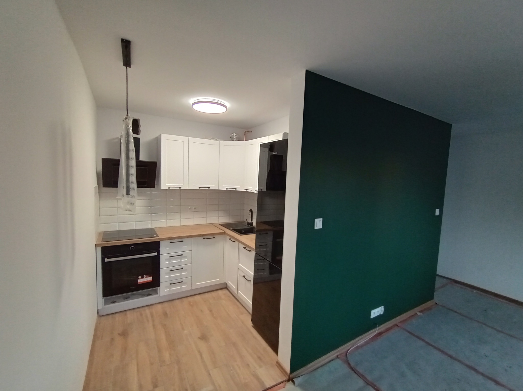 Nowoczesna, jasna kuchnia z białymi szafkami, drewnianym blatem i ciemnozieloną ścianą. Na podłodze panele imitujące drewno, a na ścianie nad blatem białe płytki. Lampa wisząca w folii ochronnej.
