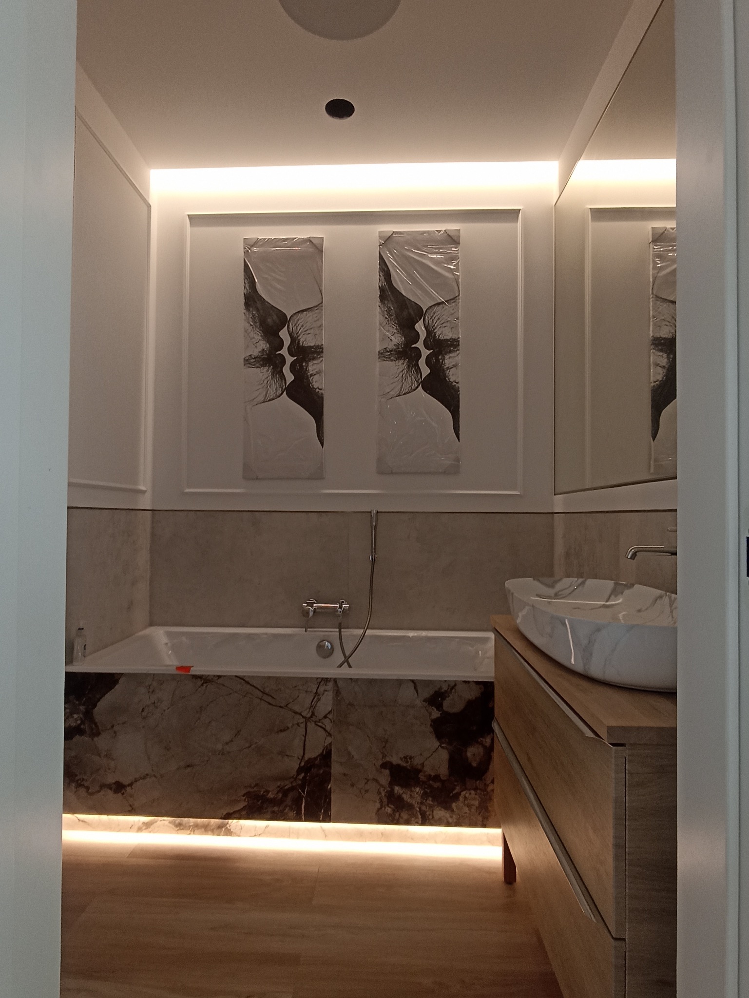 Nowoczesna łazienka z marmurową obudową wanny i umywalką, podświetleniem LED pod wanną i nad lustrem, oraz dwoma obrazami z motywem twarzy nad wanną, jeszcze zabezpieczonymi folią.