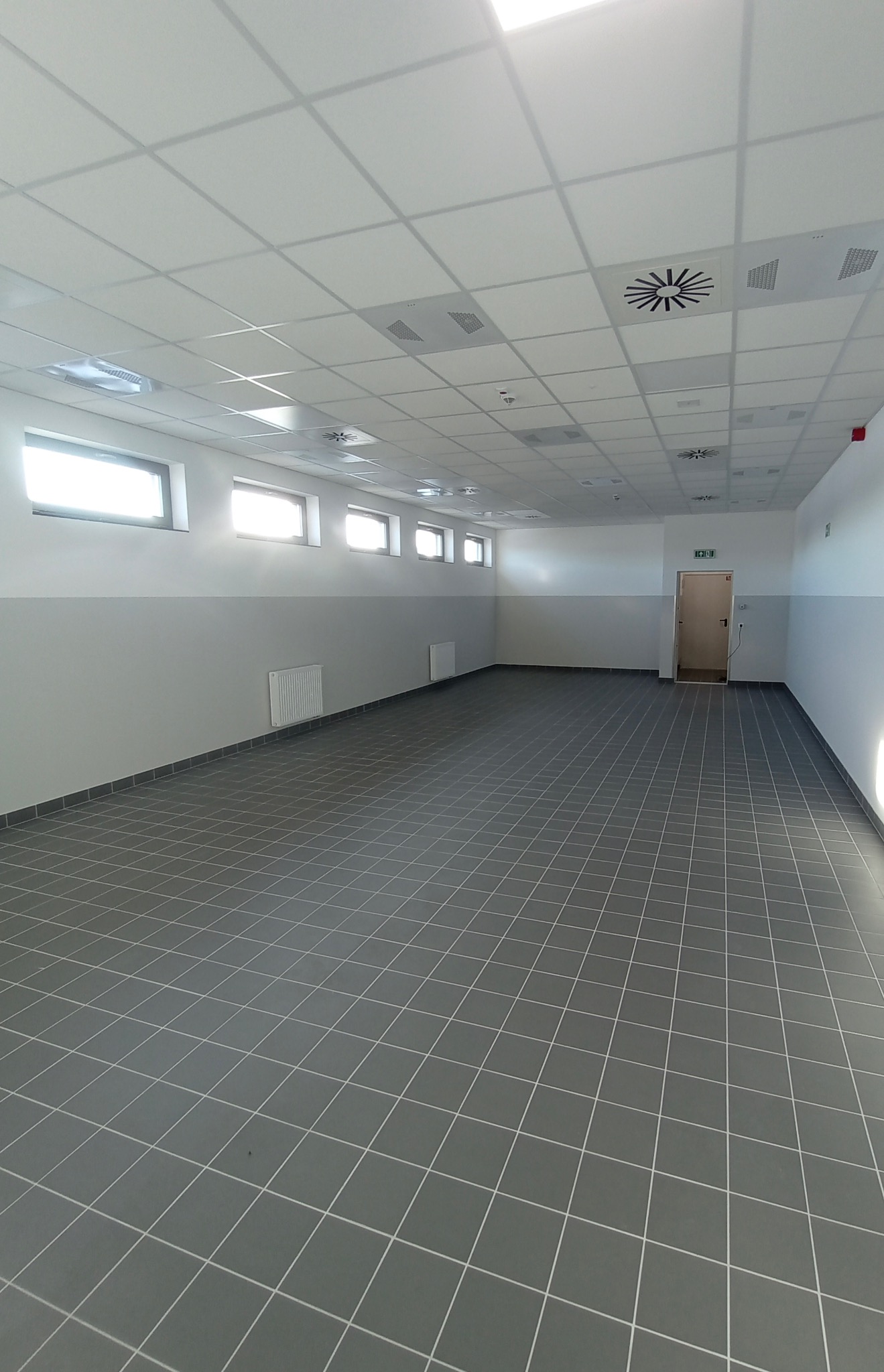 Puste pomieszczenie biurowe po remoncie z szarą podłogą w kratkę, biało-szare ściany z grzejnikami, sufitem kasetonowym z wentylacją i oknami w górnej części ściany.