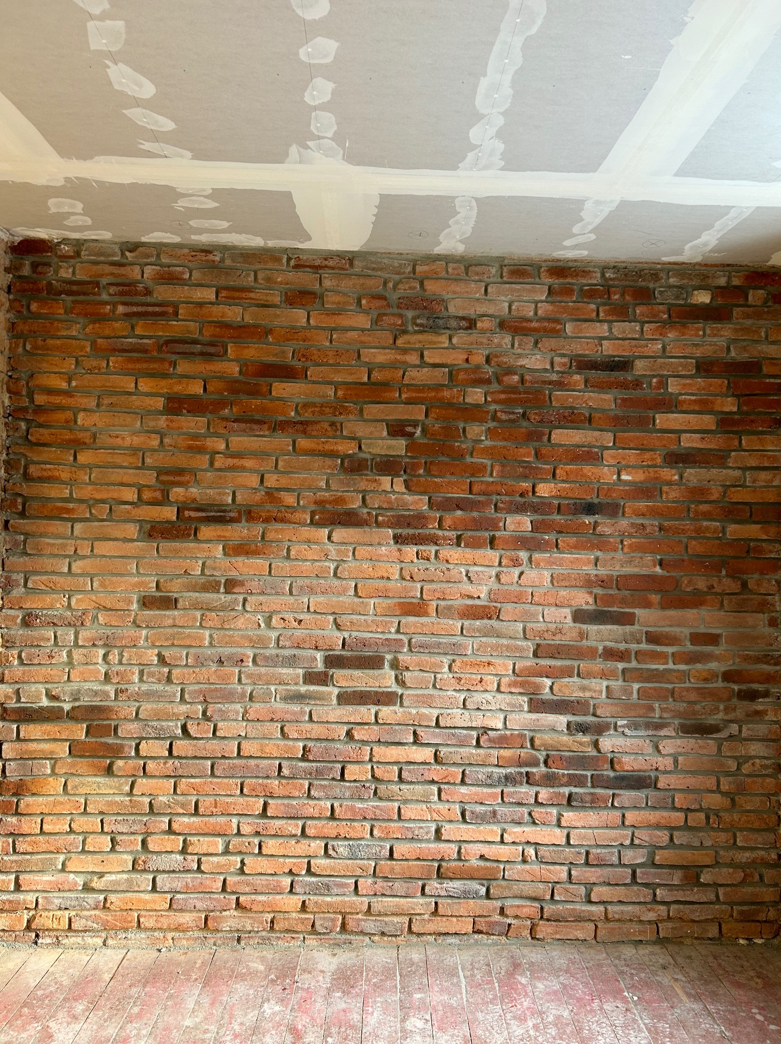 Ceglana ściana w pomieszczeniu w Legnicy, z widocznymi śladami po szpachlowaniu na suficie z płyt kartonowo-gipsowych oraz fragmentem podłogi z desek, w trakcie prac wykończeniowych.
