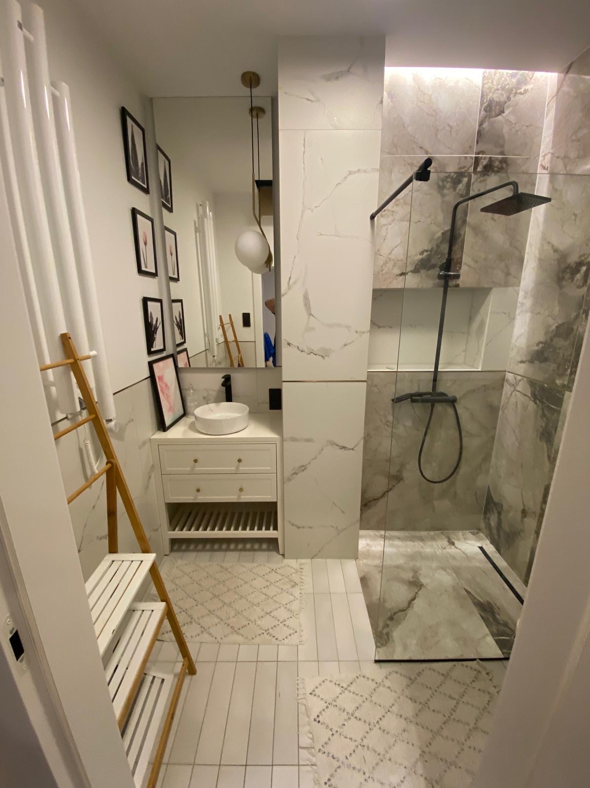 Nowoczesna łazienka z kabiną prysznicową typu walk-in wykończoną płytkami imitującymi marmur, białą szafką pod umywalkę z okrągłą nablatową umywalką, drewnianym wieszakiem na ręczniki...