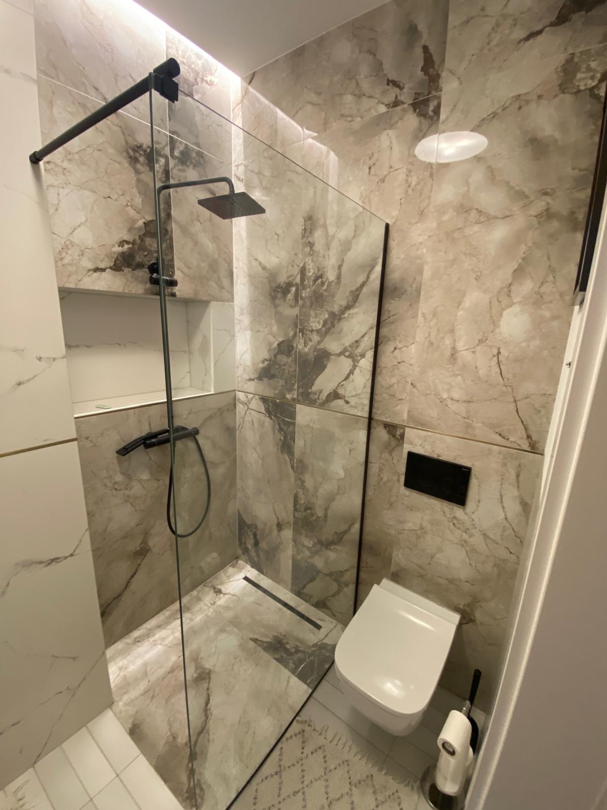 Nowoczesna łazienka z kabiną prysznicową typu walk-in z czarną armaturą, marmurowymi płytkami na ścianach i podłodze, oraz podwieszaną toaletą.