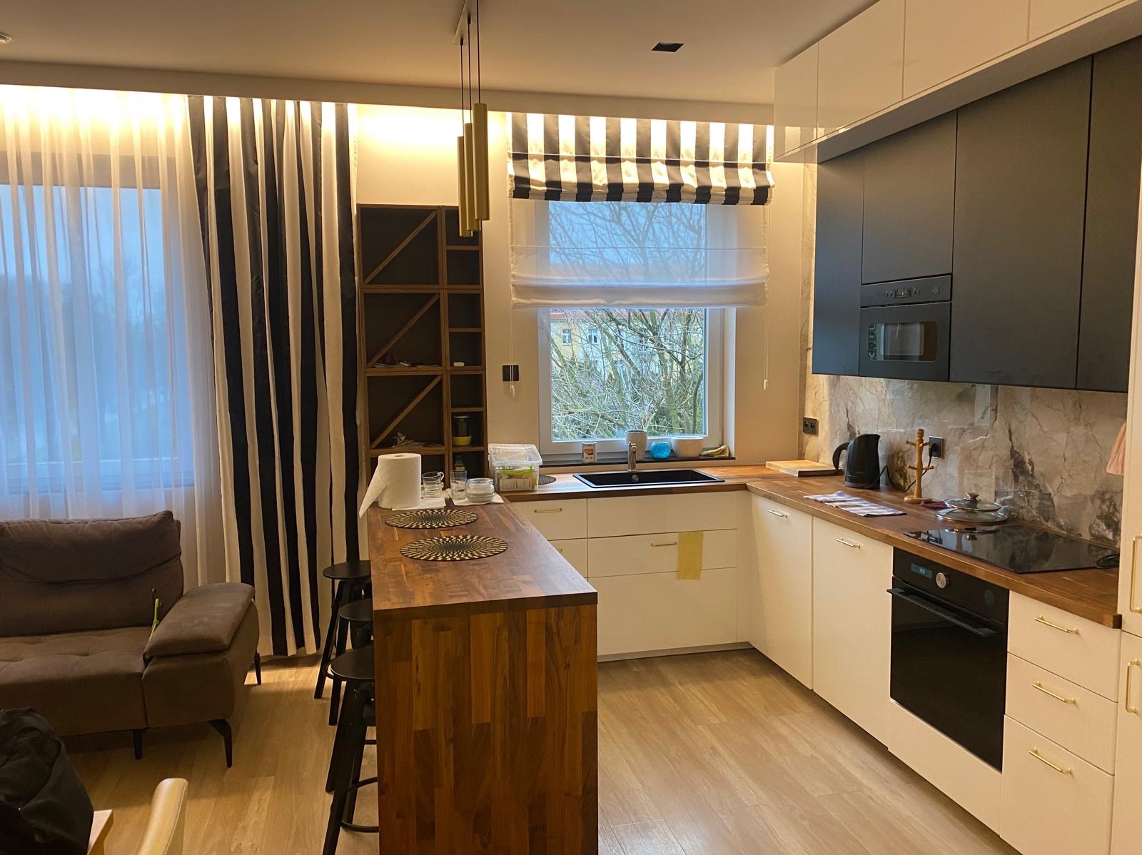 Nowoczesna kuchnia z drewnianym blatem wyspowym, czarnymi szafkami górnymi i białymi dolnymi, okno z roletą w paski, widoczny fragment salonu z sofą.
