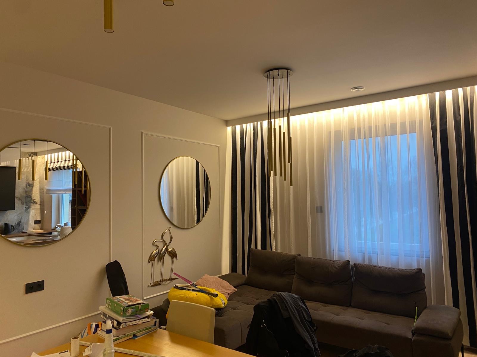 Jasne wnętrze salonu z okrągłymi lustrami w złotych ramach, brązową sofą i lampą wiszącą z złotych tub, oświetlone listwą LED pod sufitem.