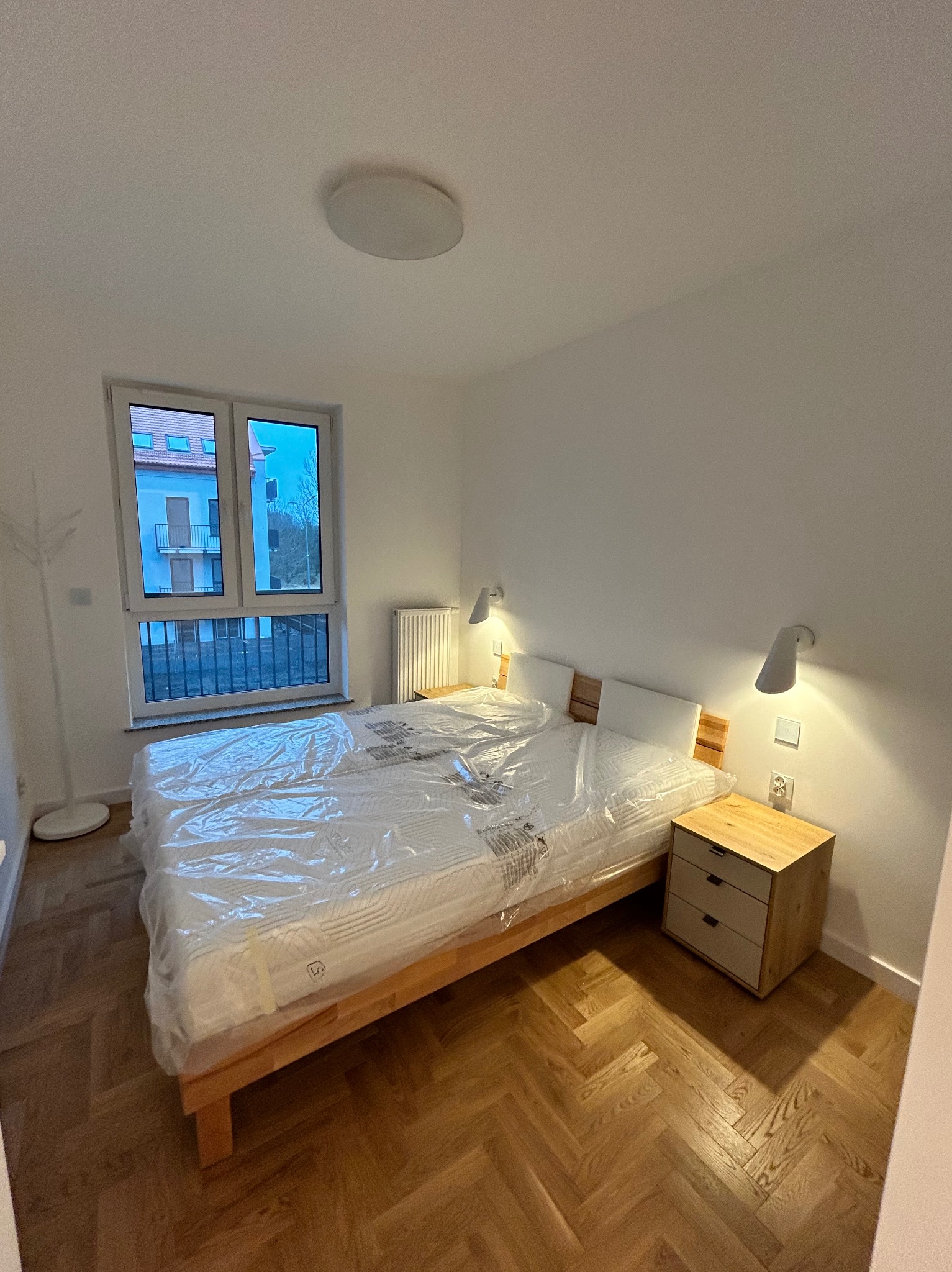 Nowe, podwójne łóżko z drewnianą ramą i materacem w folii ochronnej, ustawione w pokoju z parkietem w jodełkę, obok szafka nocna i lampki ścienne, widok przez okno na budynek.