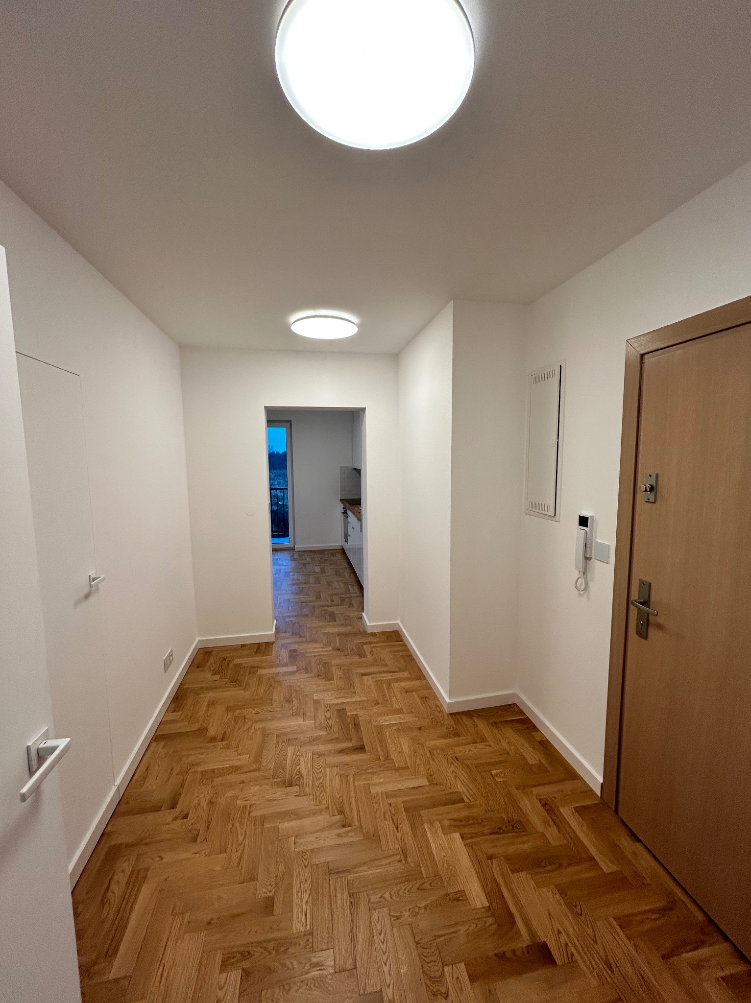 Korytarz w mieszkaniu po remoncie z parkietem ułożonym w jodełkę, pomalowane na biało ściany, widoczne drzwi wejściowe i wejście do kuchni.