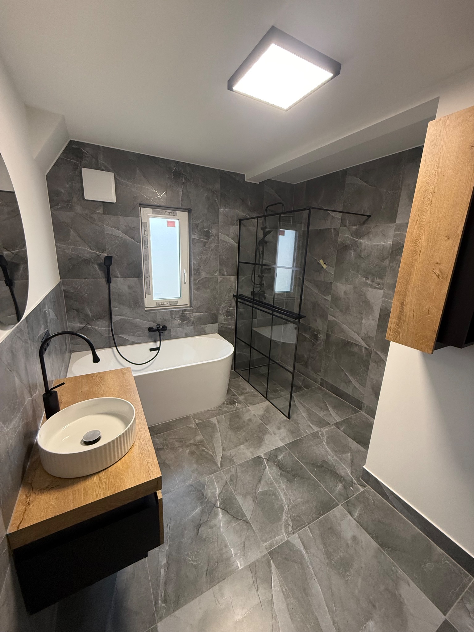 Nowoczesna aranżacja łazienki w Legnicy z szarymi płytkami imitującymi marmur, kabiną prysznicową typu walk-in z czarnymi profilami, wolnostojącą białą wanną i umywalką nablatową na drewnianym blacie.