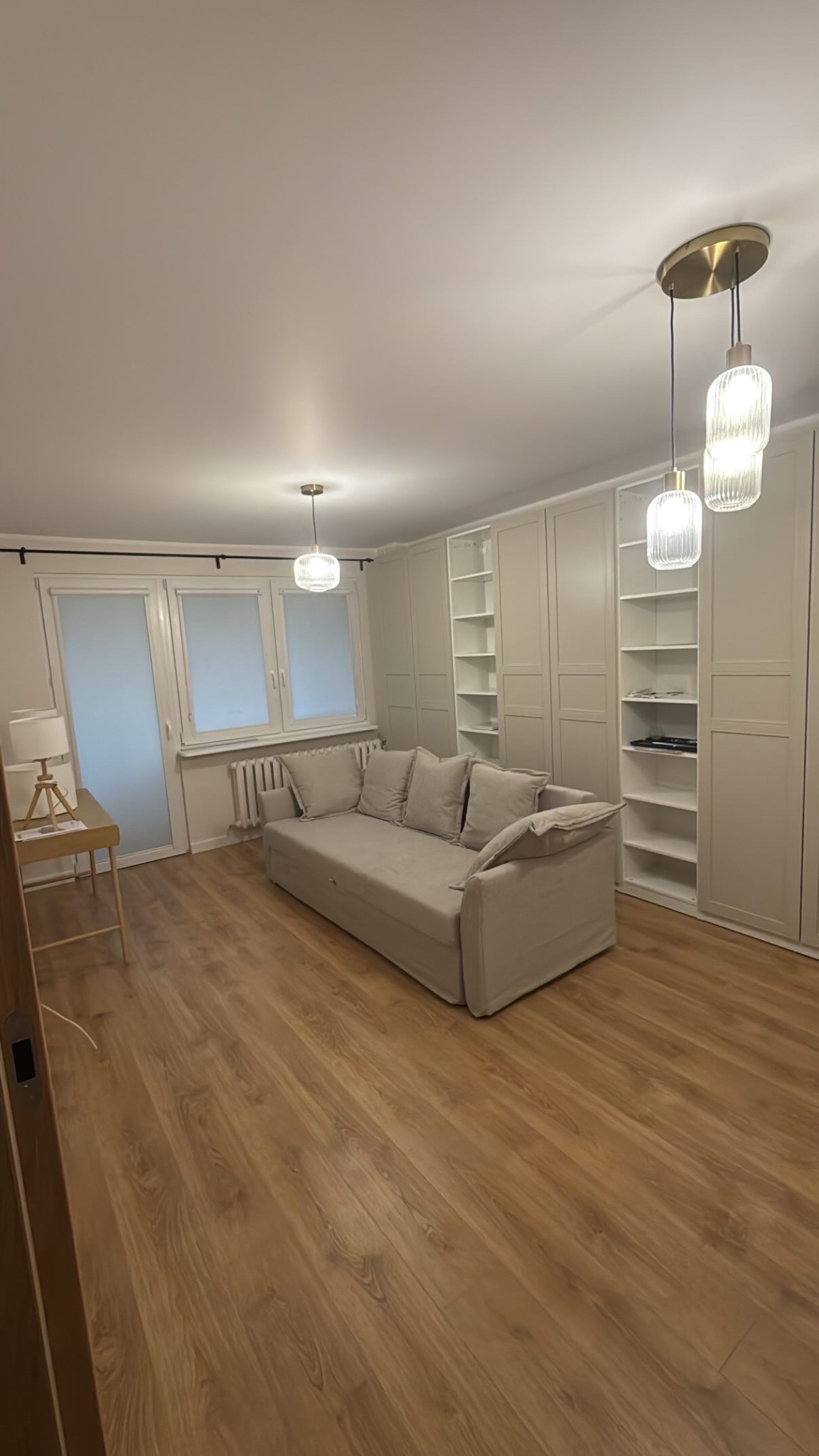 Jasne wnętrze pokoju po remoncie: drewniana podłoga, beżowa sofa z poduszkami, białe szafy w zabudowie, okno z grzejnikiem i lampy wiszące.