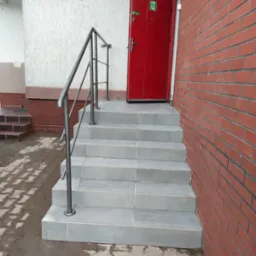 Wyłożone jasnoszarą terakotą schody z metalową barierką prowadzą do czerwonych drzwi z piktogramem ewakuacyjnym. Ceglana ściana z prawej strony.