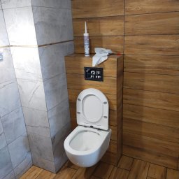 JACEK ZIELIŃSKI USŁUGI REMONTOWO-WYKOŃCZENIOWE - Wykończona toaleta podwieszana w łazience z drewnopodobnymi i marmurowymi płytkami. Widoczna silikonowa tuba i narzędzia na zabudowie.