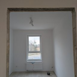 JACEK ZIELIŃSKI USŁUGI REMONTOWO-WYKOŃCZENIOWE - Wnętrze pokoju z oknem po remoncie, widoczne ślady po szpachlowaniu ościeżnicy, podłoga wyłożona panelami, białe ściany i sufit z lampą.