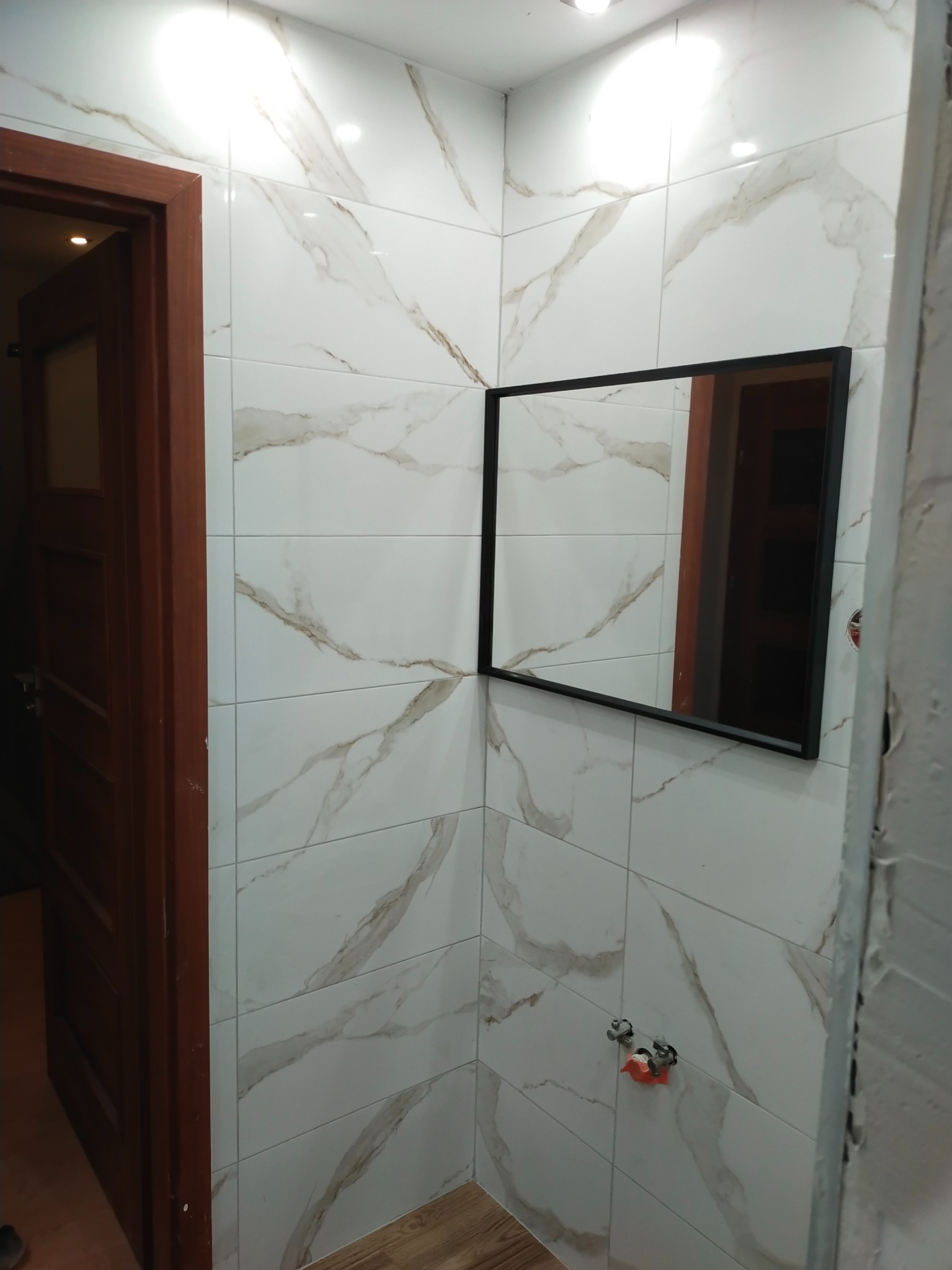 Wykończona łazienka z białymi płytkami imitującymi marmur, czarne prostokątne lustro, drewniana podłoga i częściowo widoczne zawory. Widoczny fragment niedokończonej ściany po prawej stronie.