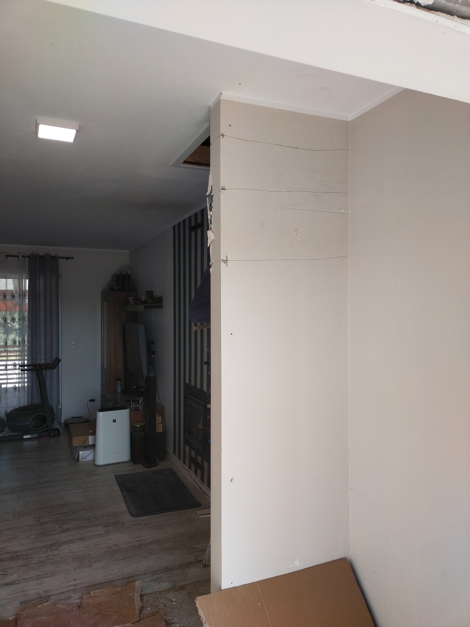 Wnętrze w trakcie prac, widoczna ściana z płyt karton-gips, szpachlowana, ale jeszcze nie pomalowana, wystające przewody, oraz fragment wykończonego pokoju z szarą tapetą w paski i sprzętem fitness...
