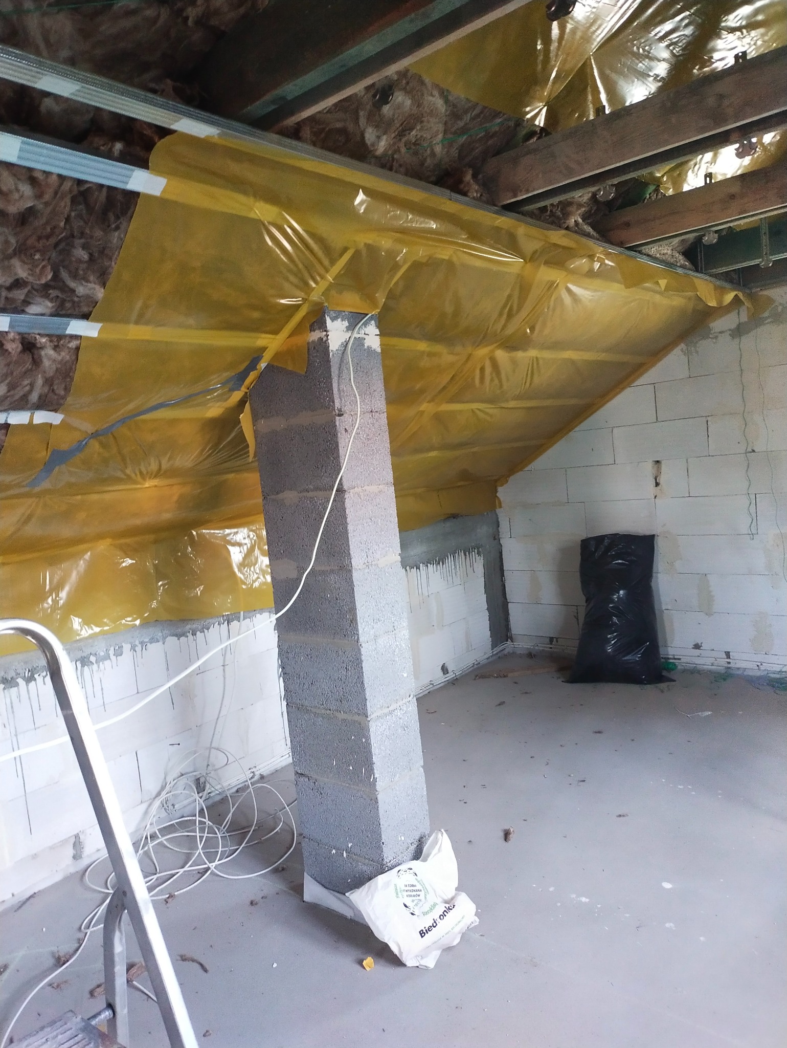 Przygotowania do wykończenia poddasza: izolacja z folią paroizolacyjną, murowany filar z betonu komórkowego z poprowadzoną instalacją elektryczną, widoczna drabina i worek na śmieci.