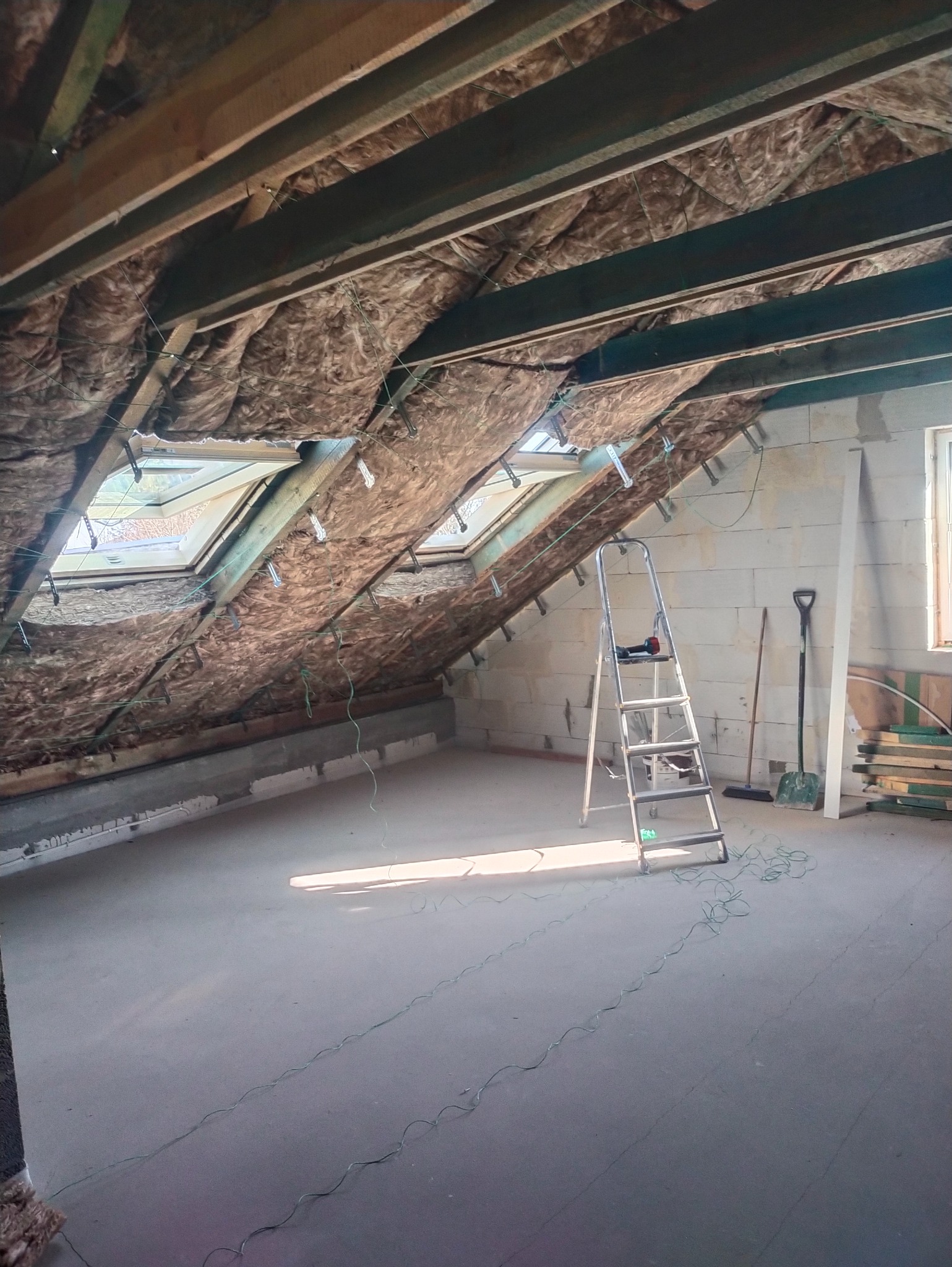 Poddasze z oknami dachowymi w trakcie izolacji, widoczne drewniane belki, wełna mineralna, drabina aluminiowa, narzędzia i materiały budowlane.