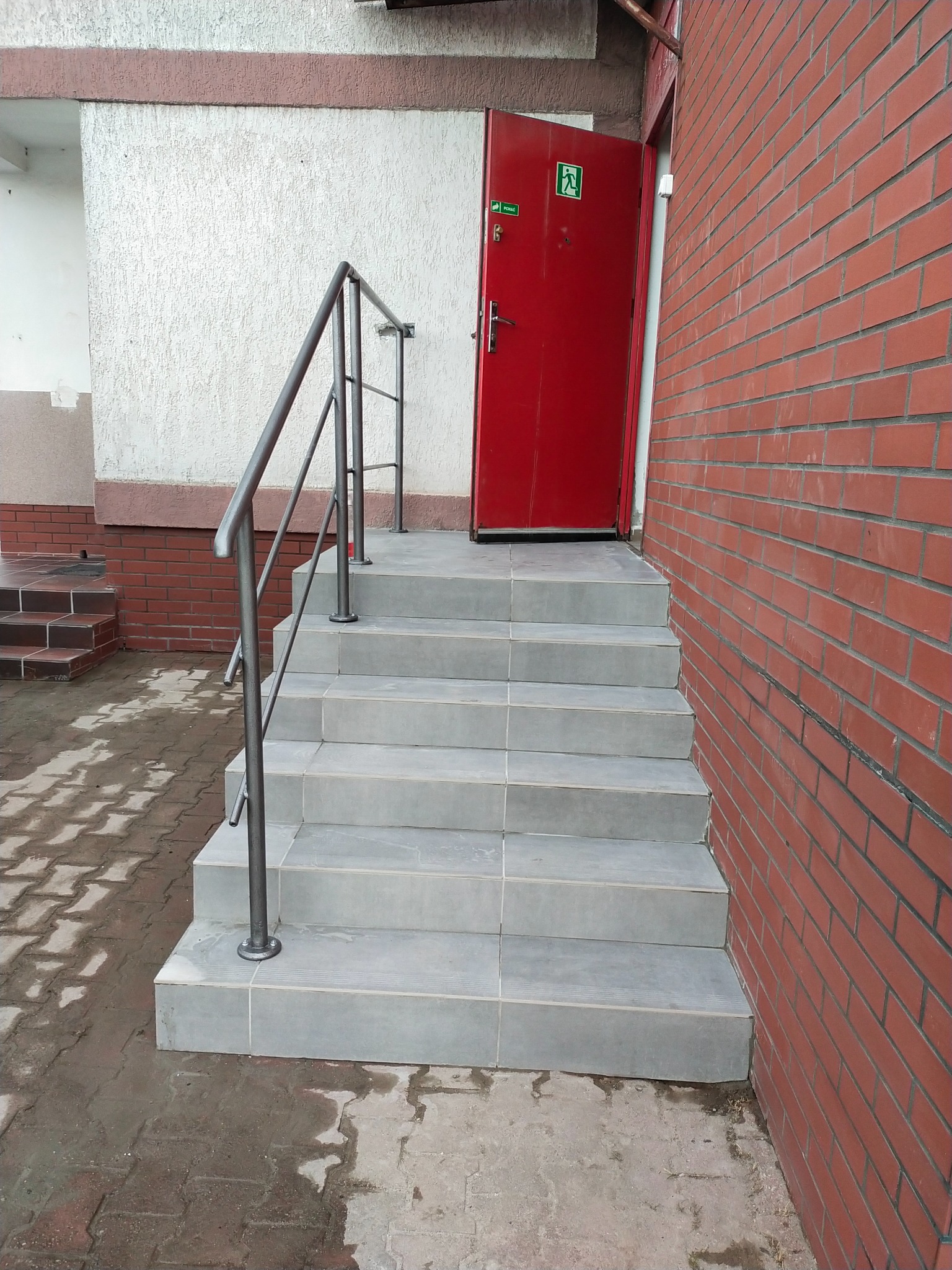 Wyłożone jasnoszarą terakotą schody z metalową barierką prowadzą do czerwonych drzwi z piktogramem ewakuacyjnym. Ceglana ściana z prawej strony.