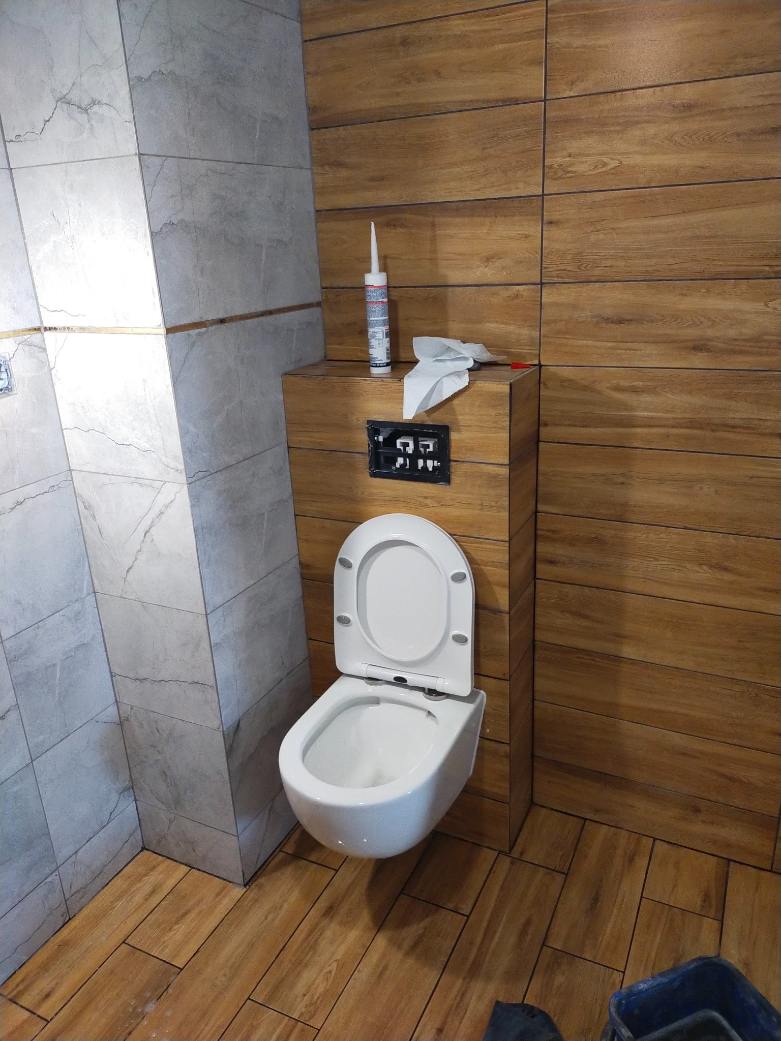 Wykończona toaleta podwieszana w łazience z drewnopodobnymi i marmurowymi płytkami. Widoczna silikonowa tuba i narzędzia na zabudowie.