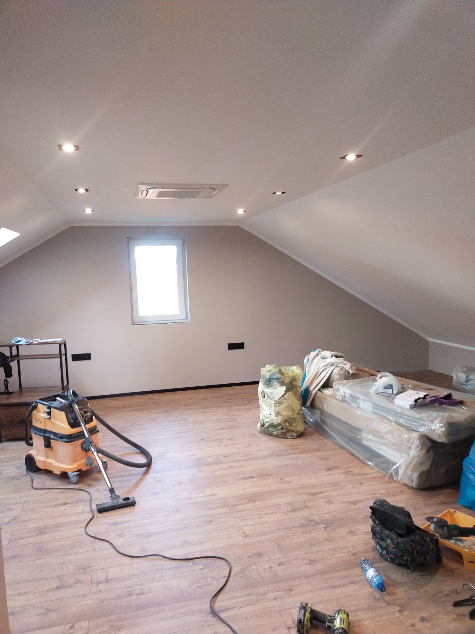 Poddasze po remoncie z drewnopodobną podłogą, świeżo pomalowanymi ścianami i zamontowanym oświetleniem. Widoczny odkurzacz przemysłowy i narzędzia.