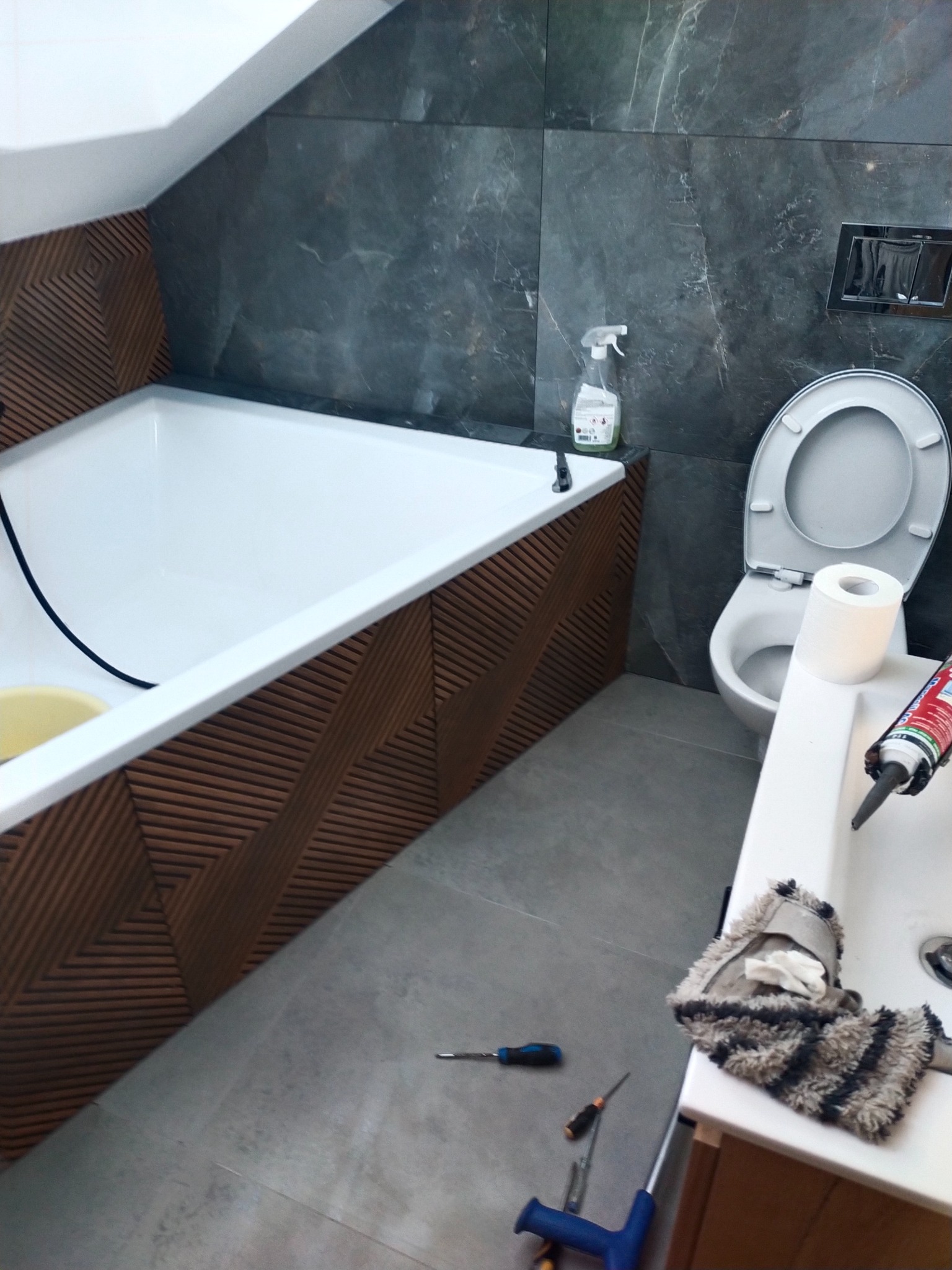 Wykończona łazienka z wanną w zabudowie z drewnopodobnymi panelami, szarą glazurą, toaletą i umywalką. Widoczne narzędzia sugerują prace wykończeniowe.