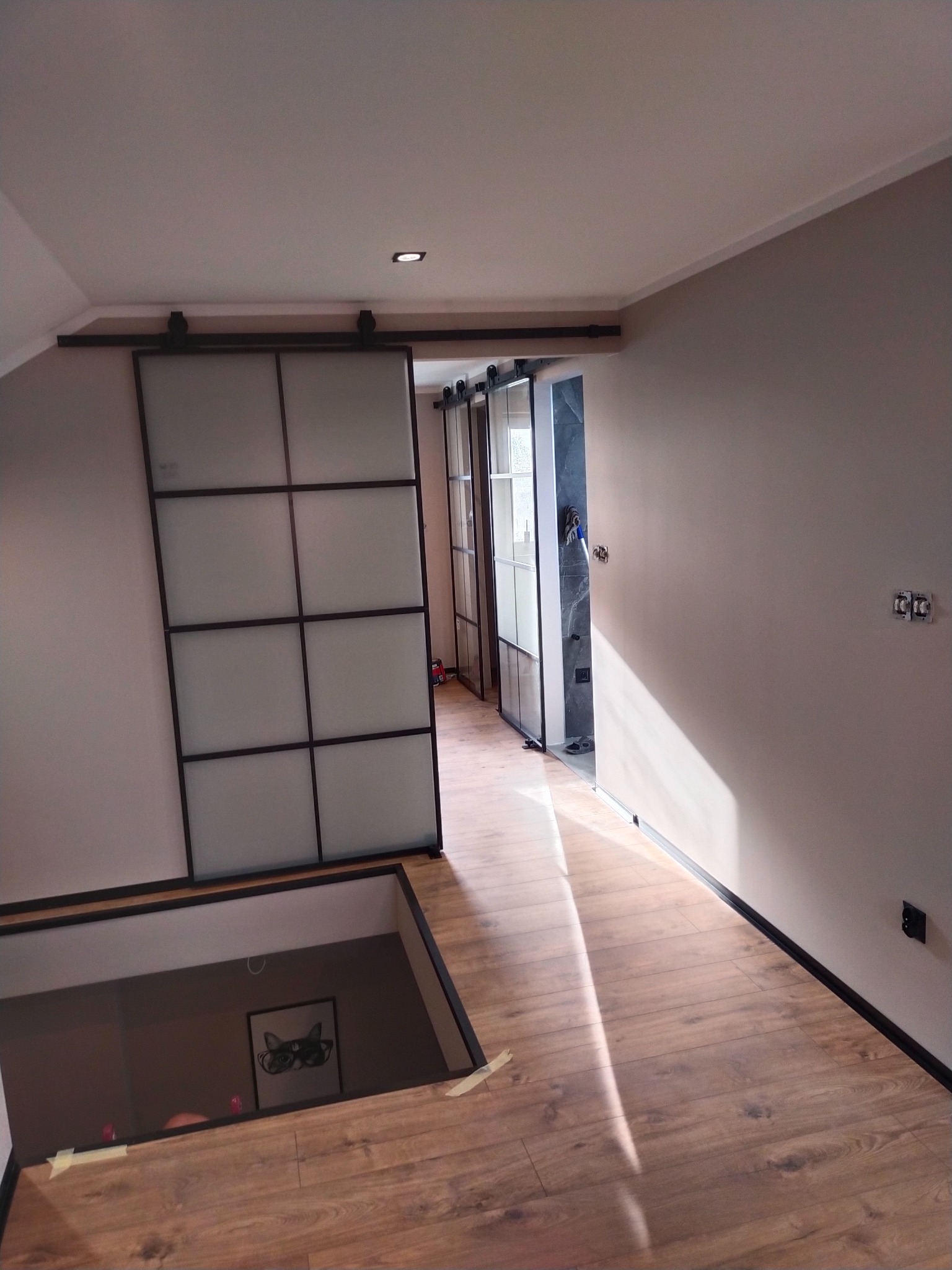 Loftowe drzwi przesuwne z matowego szkła na poddaszu, widok na łazienkę z szarą glazurą i drewnianą podłogę. Minimalistyczny design, ciepłe oświetlenie.