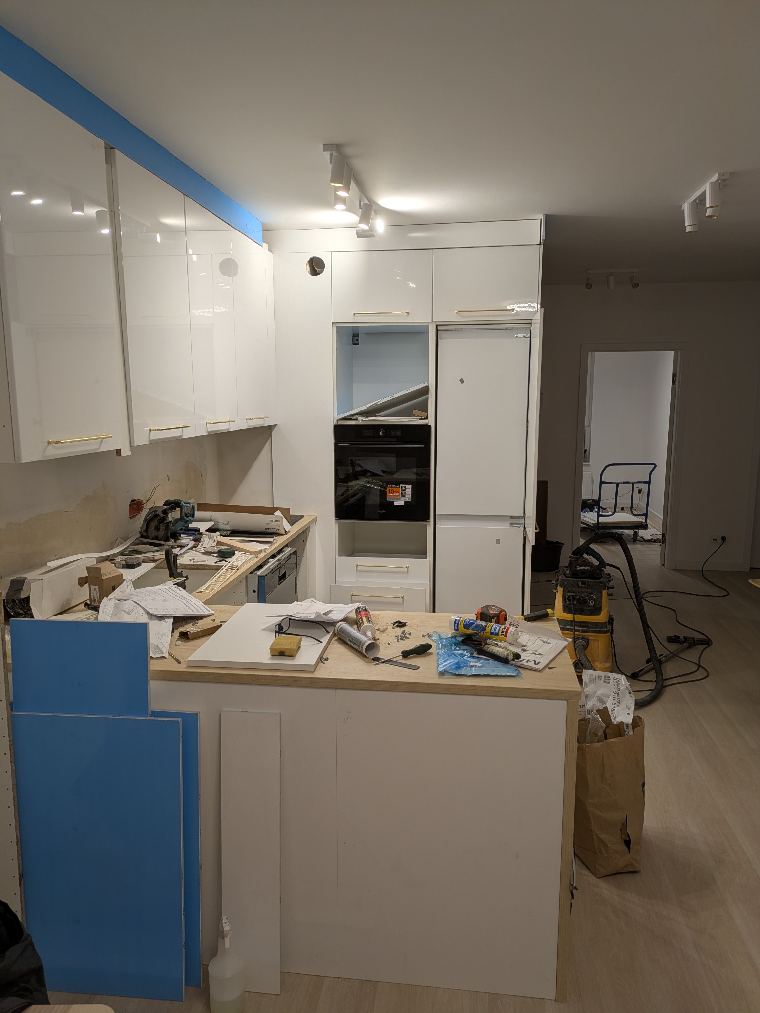 Wnętrze kuchni w trakcie montażu: białe szafki z złotymi uchwytami, sprzęt AGD w zabudowie, narzędzia na blacie, niebieskie fronty czekające na montaż, odkurzacz przemysłowy.