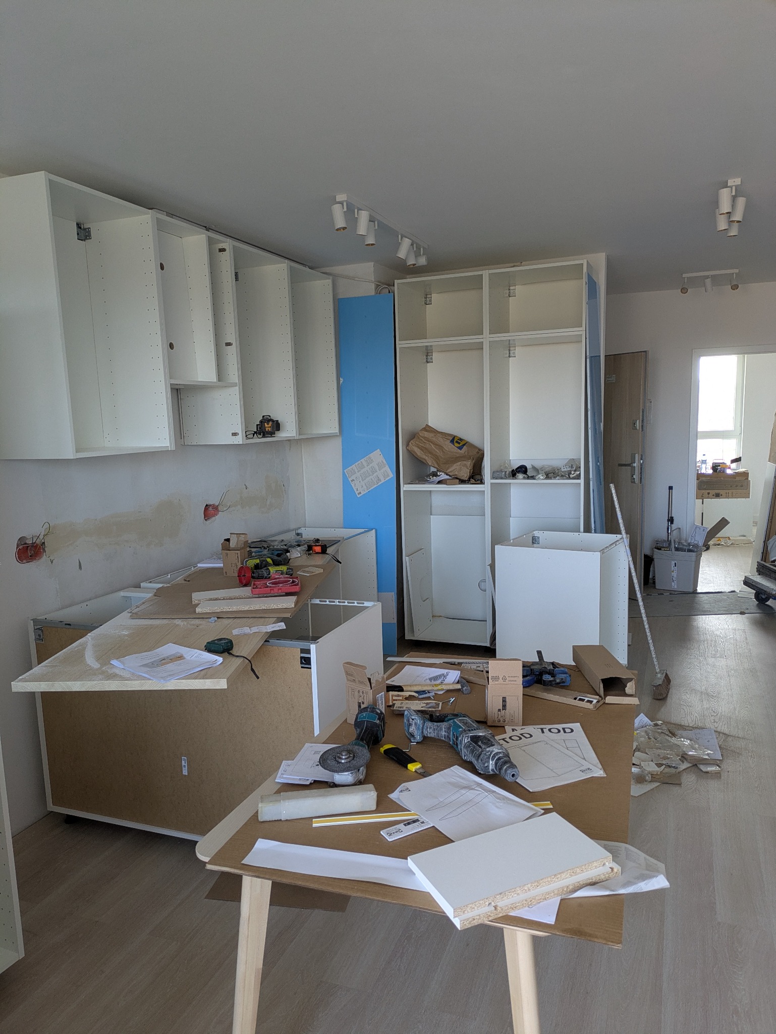Montaż kuchni w trakcie: widoczne niedokończone szafki wiszące i stojące, narzędzia na blacie, elementy instalacji elektrycznej. W tle drzwi i okno.