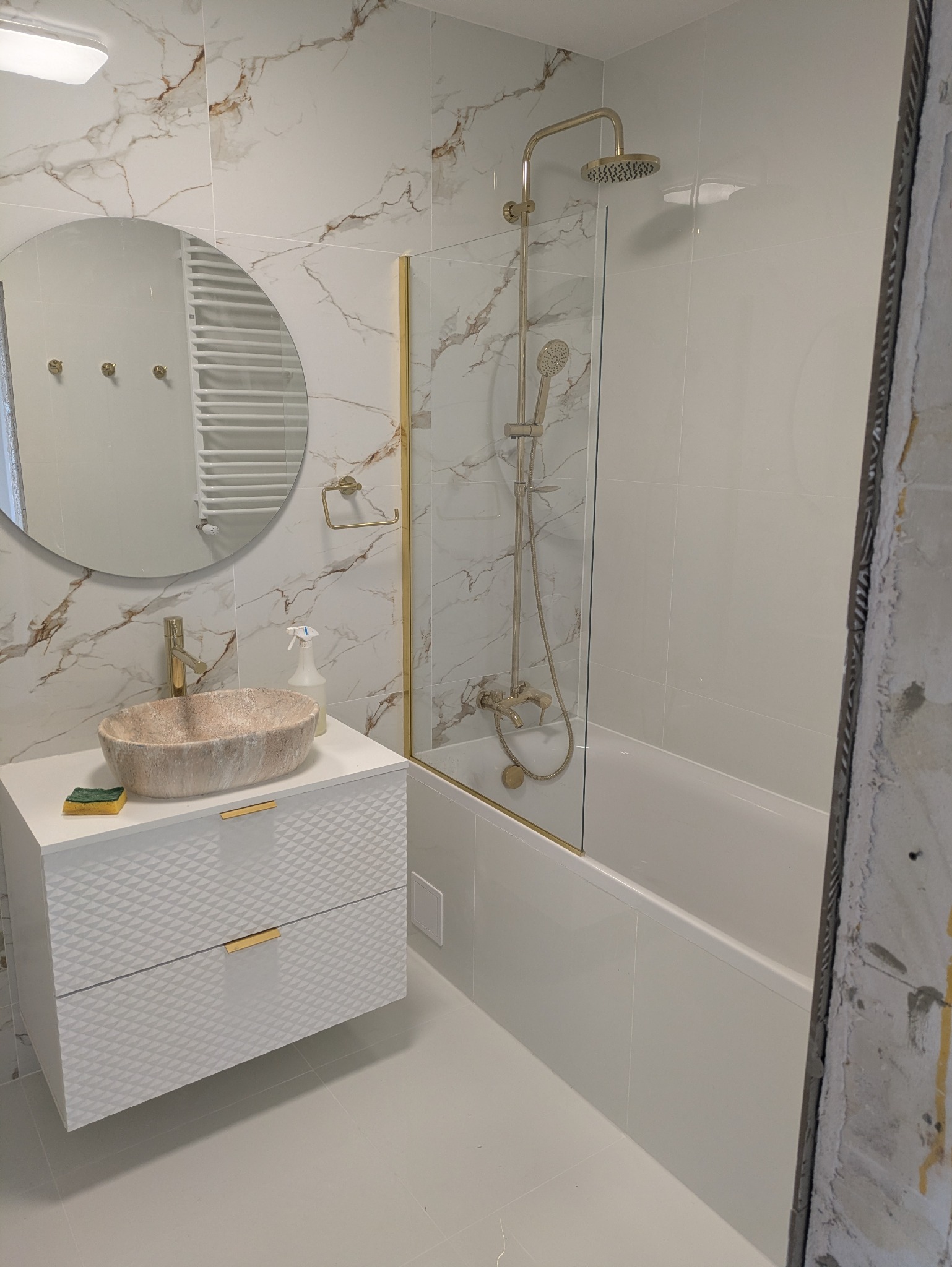 Wykończona łazienka z marmurowymi płytkami, okrągłym lustrem i umywalką w odcieniach beżu. Kabina prysznicowa ze złotymi detalami i biała szafka pod umywalką.
