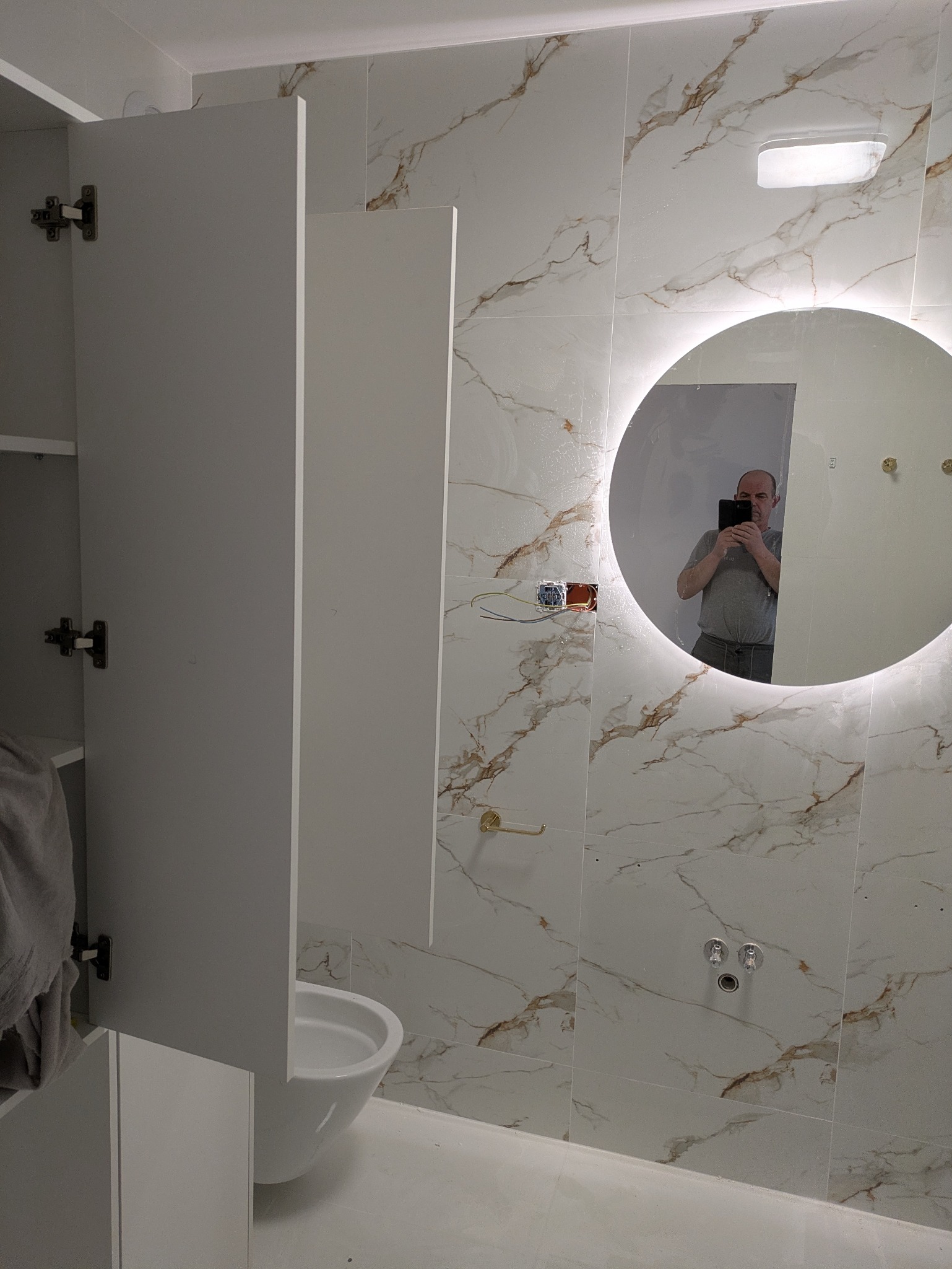 Biała łazienka z marmurowymi płytkami, okrągłym lustrem z podświetleniem LED i otwartą szafką. Widoczny fragment toalety i instalacja elektryczna.