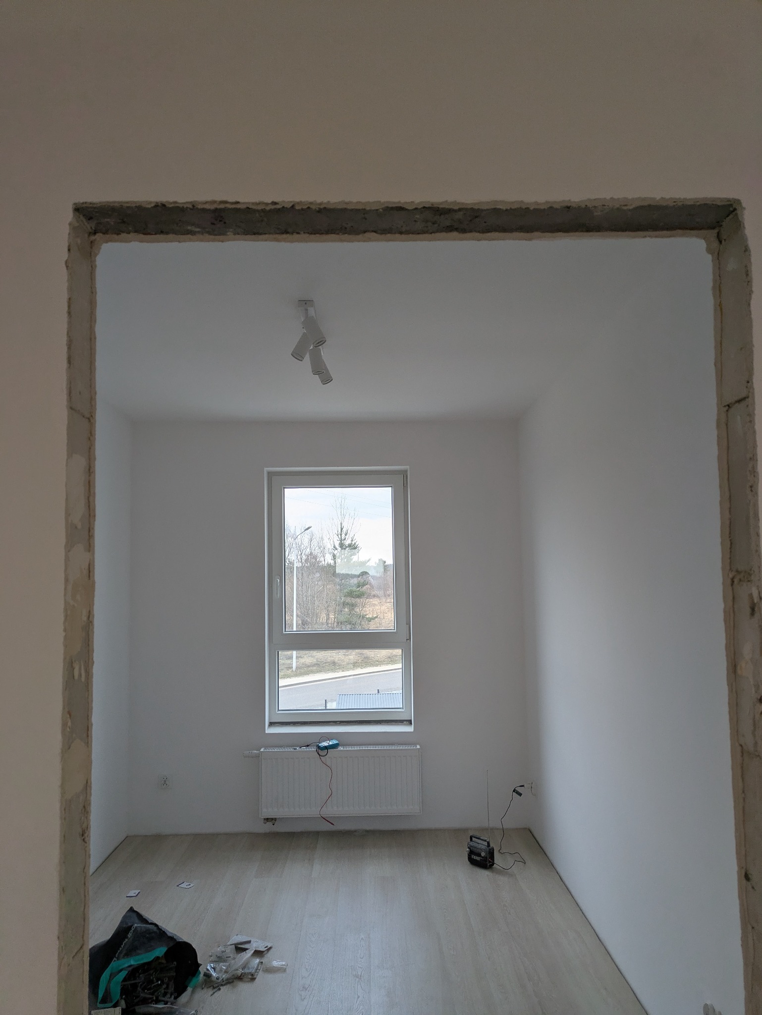Wnętrze pokoju z oknem po remoncie, widoczne ślady po szpachlowaniu ościeżnicy, podłoga wyłożona panelami, białe ściany i sufit z lampą.