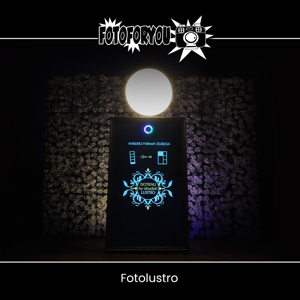 Fotolustro z podświetlanym panelem dotykowym na tle ściany z białych kwiatów, oświetlone okrągłym światłem górnym, logo 'FOTOFORYOU' w górnej części kadru.