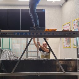 GLAZURA USŁUGI REMONTOWO-BUDOWLANE TOMASZ SZWEDO - Pracownik na rusztowaniu w lokalu gastronomicznym, widoczne zabezpieczone folią meble, plakaty na ścianie i kamery monitoringu.