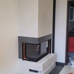 Wnękowy kominek narożny z szarą obudową, wykończony gładzią w kolorze złamanej bieli, z widocznym fragmentem pomieszczenia w tle.