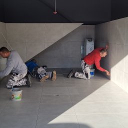 GLAZURA USŁUGI REMONTOWO-BUDOWLANE TOMASZ SZWEDO - Trzech mężczyzn w Domostawie, na kolanach, precyzyjnie wypełnia fugi pomiędzy szarymi płytkami na ścianie i podłodze, widoczny sprzęt do uzdatniania wody.