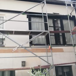 GLAZURA USŁUGI REMONTOWO-BUDOWLANE TOMASZ SZWEDO - Elewacja budynku w trakcie malowania, widoczne rusztowanie, świeżo pomalowane fragmenty ściany, czarne ramy okienne.
