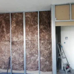 Ściana w trakcie izolacji wełną mineralną, widoczne metalowe profile konstrukcyjne, drabina oparta o ścianę, przygotowania do montażu płyty karton-gips.