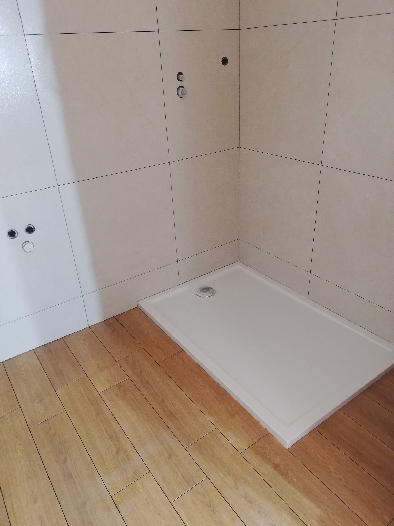 Narożnik łazienki w trakcie prac wykończeniowych: ułożona jasna, kwadratowa glazura ścienna z widocznymi otworami na armaturę, biały brodzik bez kabiny oraz podłoga wyłożona panelami imitującymi...