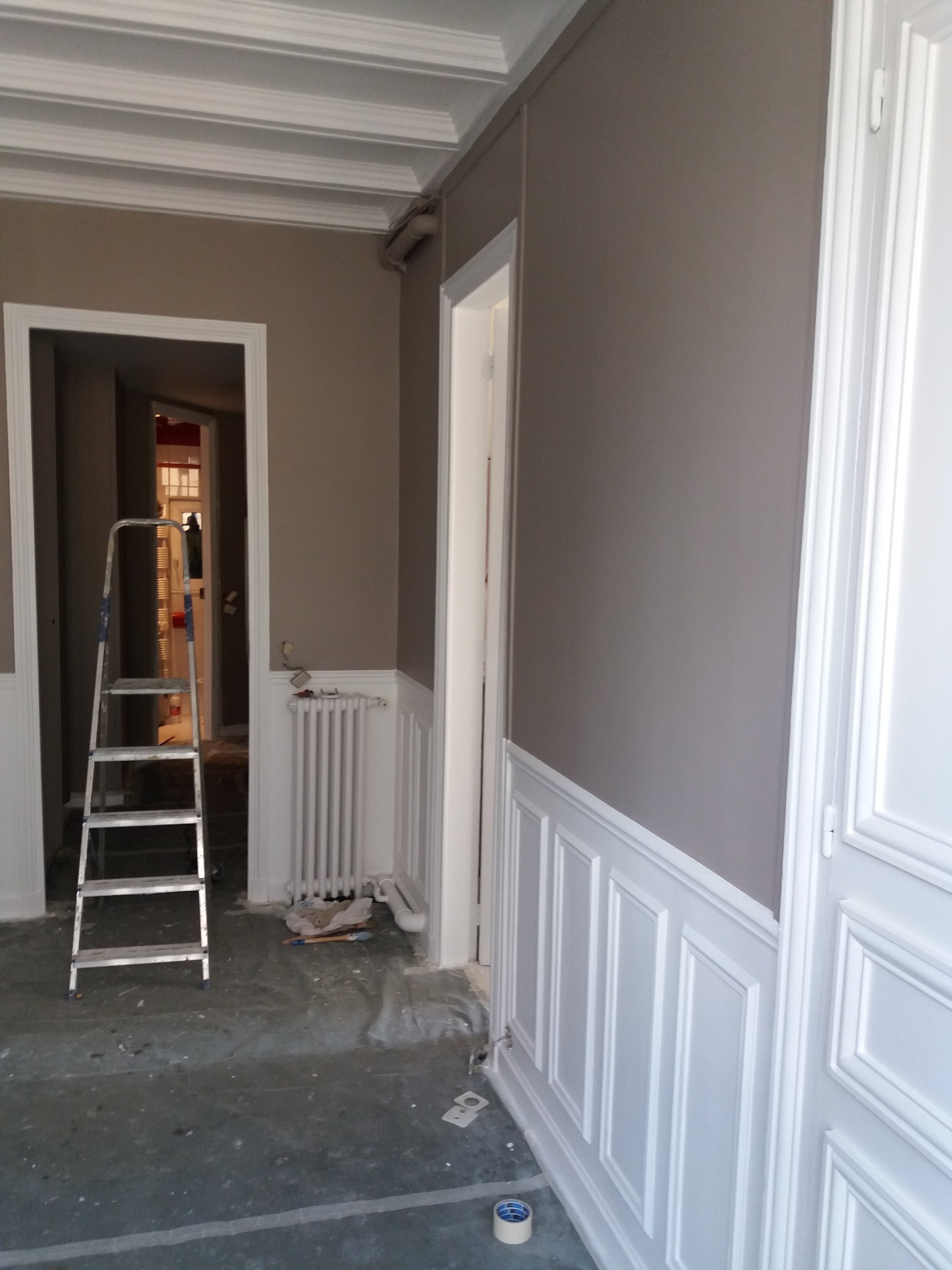 Remontowane wnętrze: szara ściana z białą boazerią, drabina malarska, grzejnik i akcesoria malarskie na zabezpieczonej podłodze.